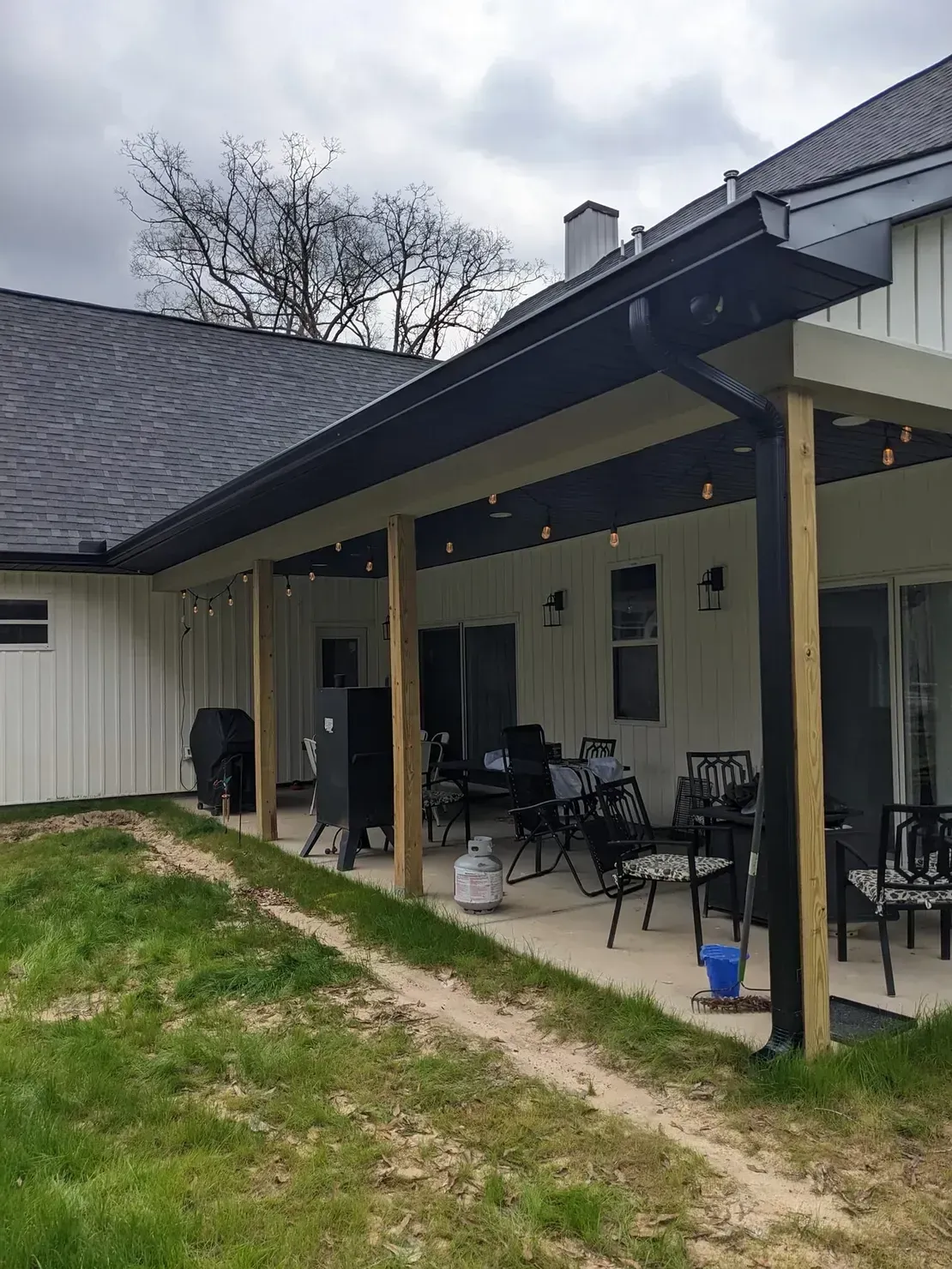 A covered patio attached to a house with vertical white siding, wooden support beams, black trim, and outdoor seating.