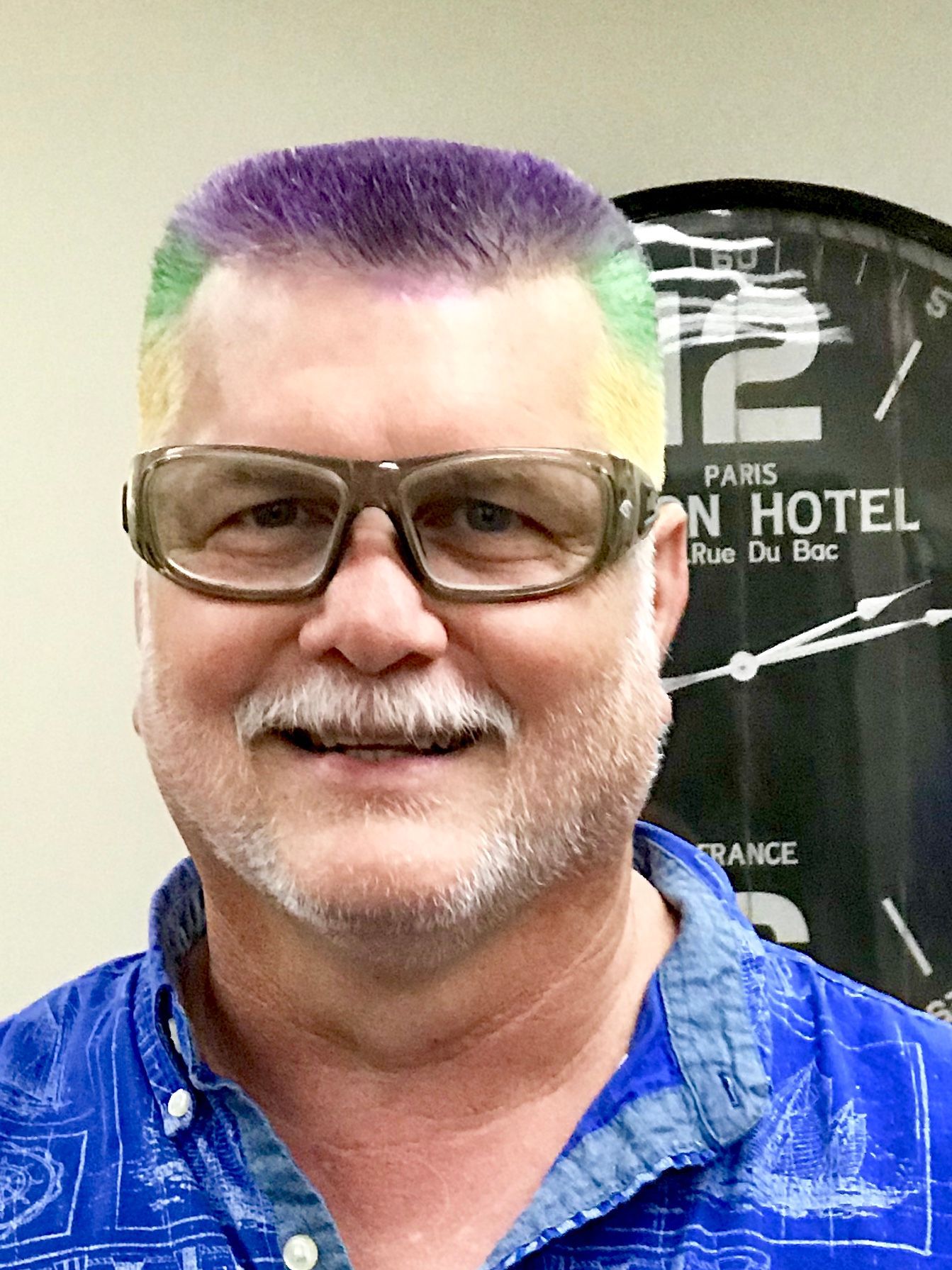 An older gentleman with glasses that has tri-colored hair for Mardi Gras. A royal purple at the top of his head, a medium to bright green at the temples, and yellow on the sides.