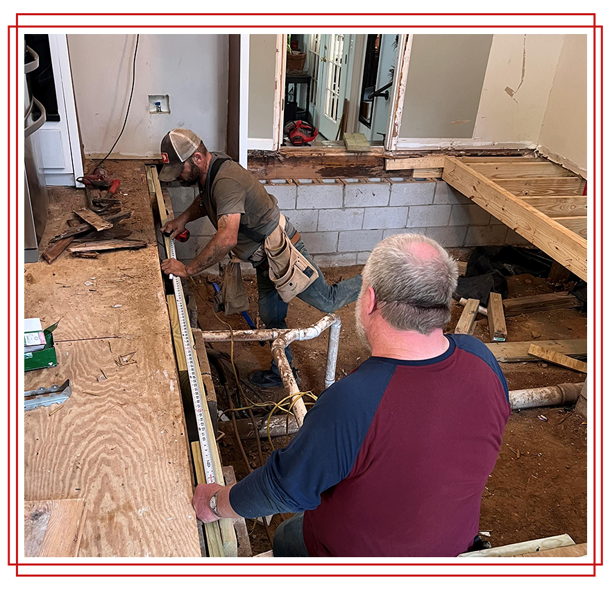 Two men repairing floor in a room with exposed foundation. One kneels, measuring, and the other holds a board.