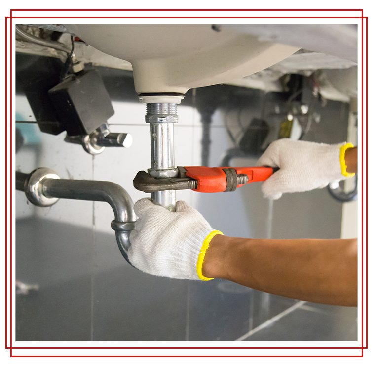 A plumber, wearing gloves, uses a wrench to tighten a pipe under a bathroom sink.