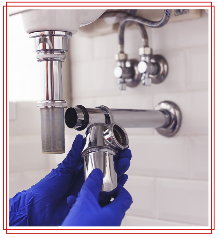 Hands in blue gloves assembling a shiny chrome sink drain trap under a white sink.