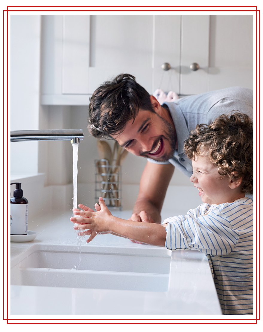 Father and child washing hands at sink, smiling. White kitchen.