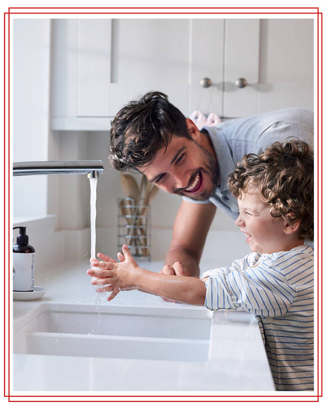 Father and child washing hands at a sink; both are smiling, in a bright kitchen.