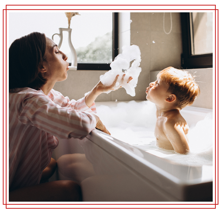 Woman and child in a bathtub playing with foam. Woman holds out foam, child looks on in a bathroom.