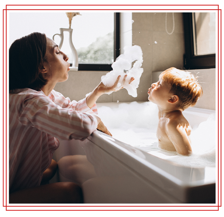 Woman in pink pjs gives bubbly foam to a toddler in a bathtub with a sunny window view.