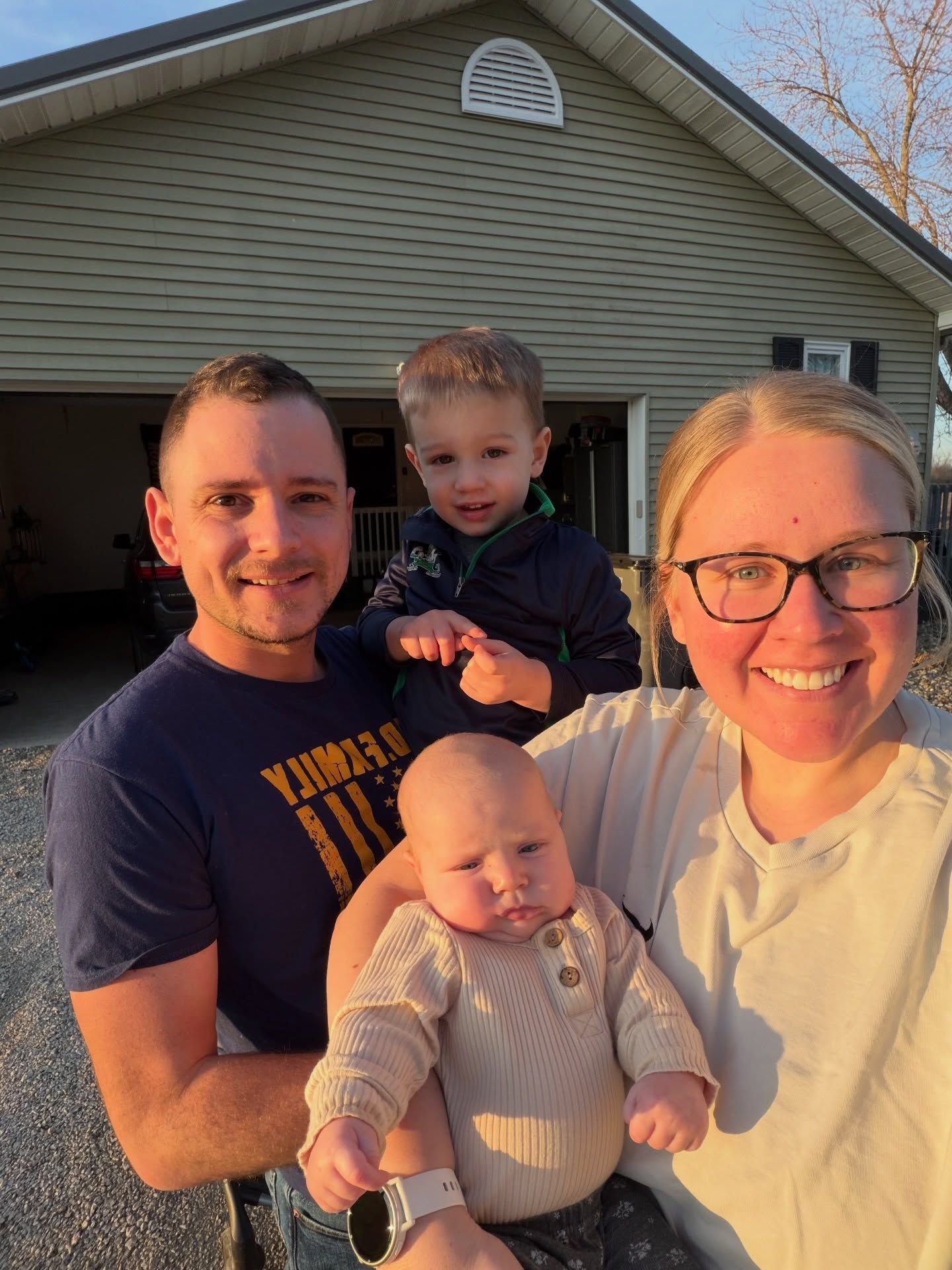 Family of four posing outside a garage, smiling and holding a baby in the foreground.