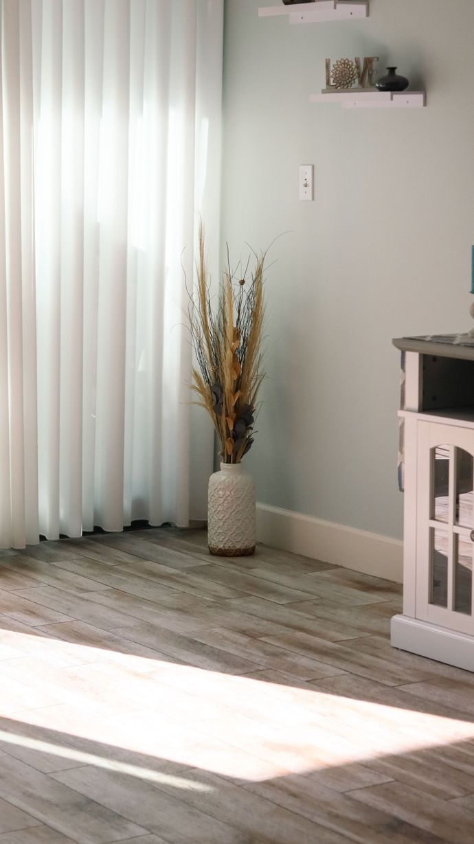 Sunlit room corner with sheer curtains, vase of dried flowers, wood floor, and white crib