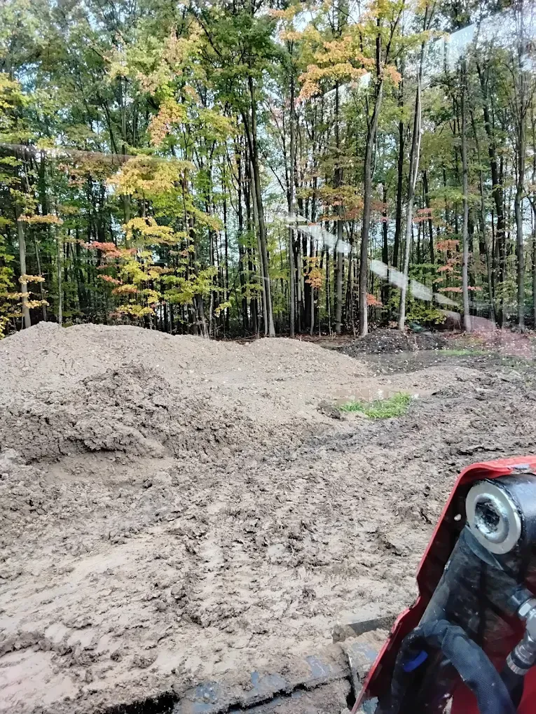 Pile of dirt in front of trees. Construction site with fall foliage. Red machinery in foreground.