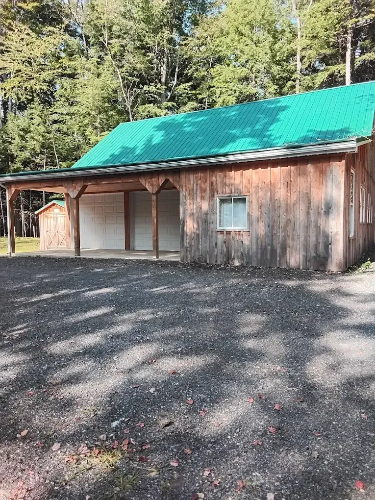 Wooden building with green roof, open carport, small window, gravel drive, and trees in background.