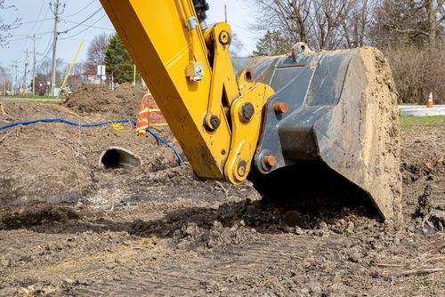Yellow excavator bucket scoops dirt from a construction site, near a culvert.