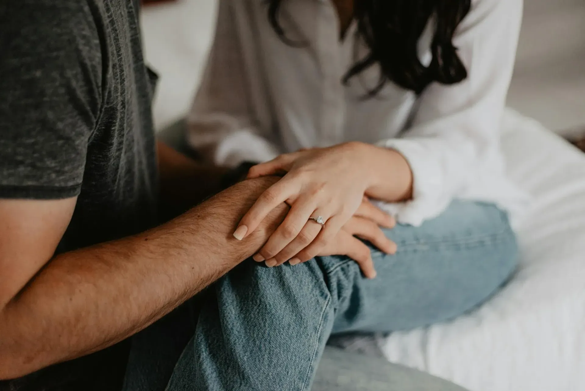 A man and a woman are sitting on a bed holding hands . the woman is wearing a wedding ring.