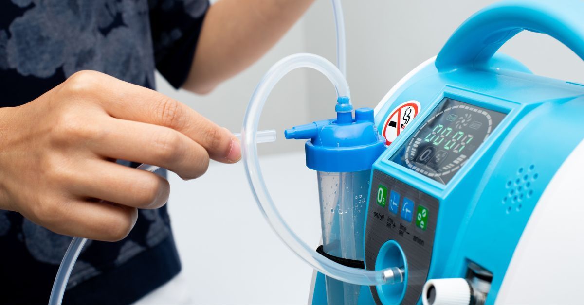 A person’s hands connecting plastic tubing to a blue portable oxygen concentrator equipped with a water-filled humidifier bottle and a