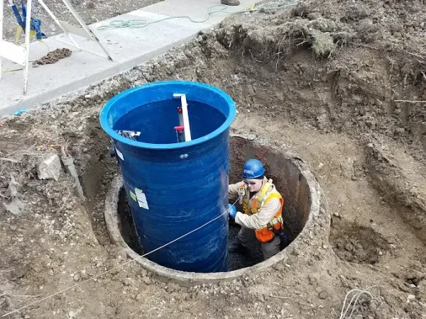 A worker in a safety vest and hard hat installs a blue cylindrical utility tank inside an open excavation pit.