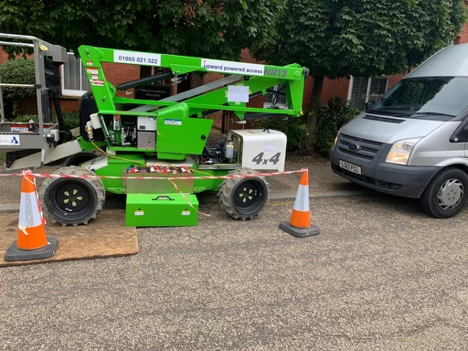 Green cherry picker and silver van parked on a paved road, orange traffic cones.