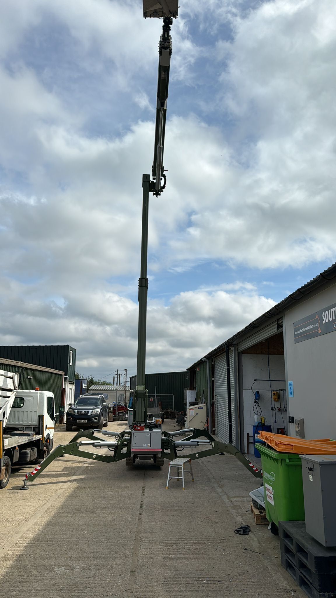 An extended aerial lift is raised high in a gravel lot next to a white building under a cloudy sky.
