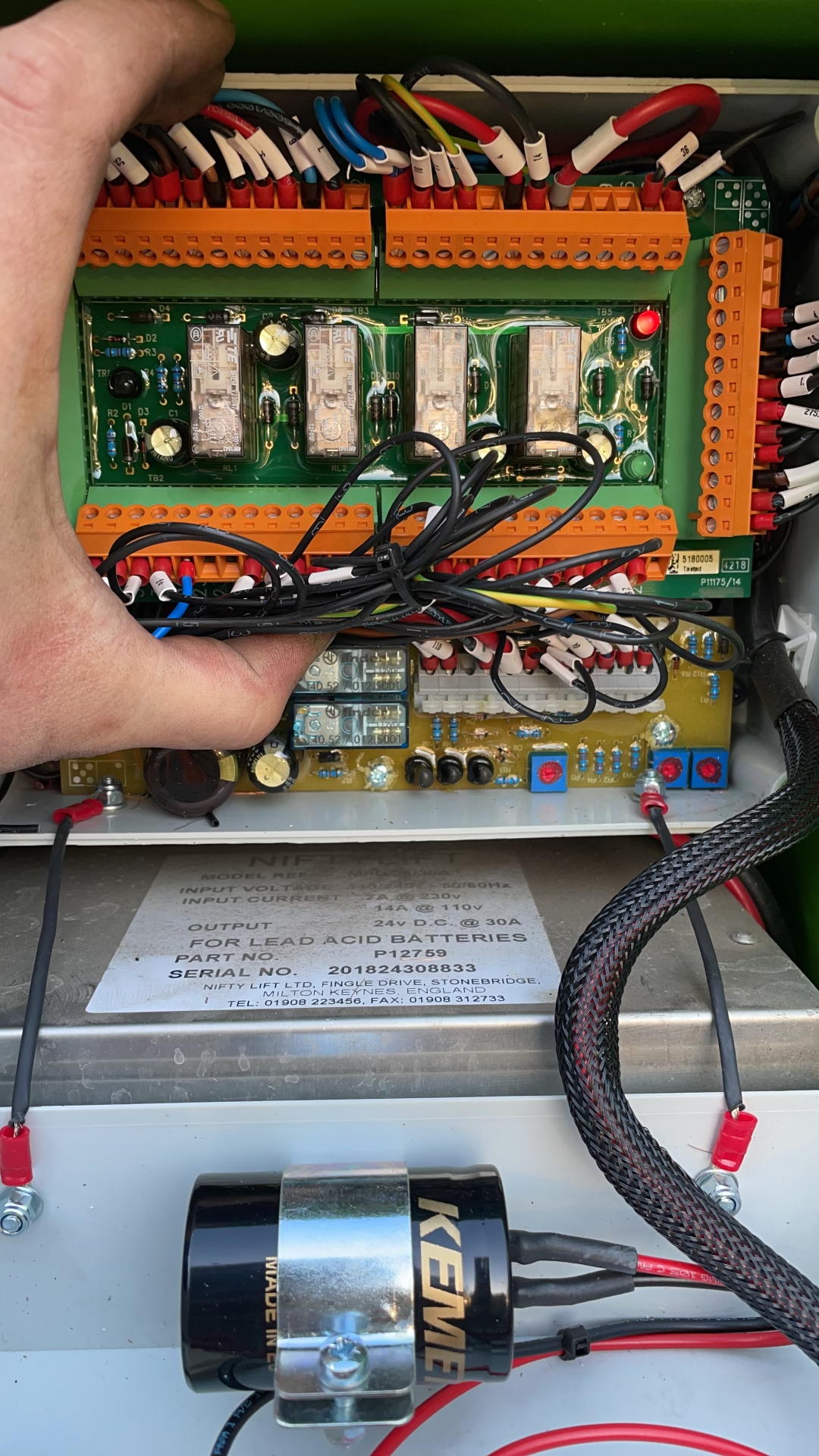 Hand touching a circuit board in an electrical panel. Wires and terminal blocks are visible. A black KEWEL solenoid is at the bottom.