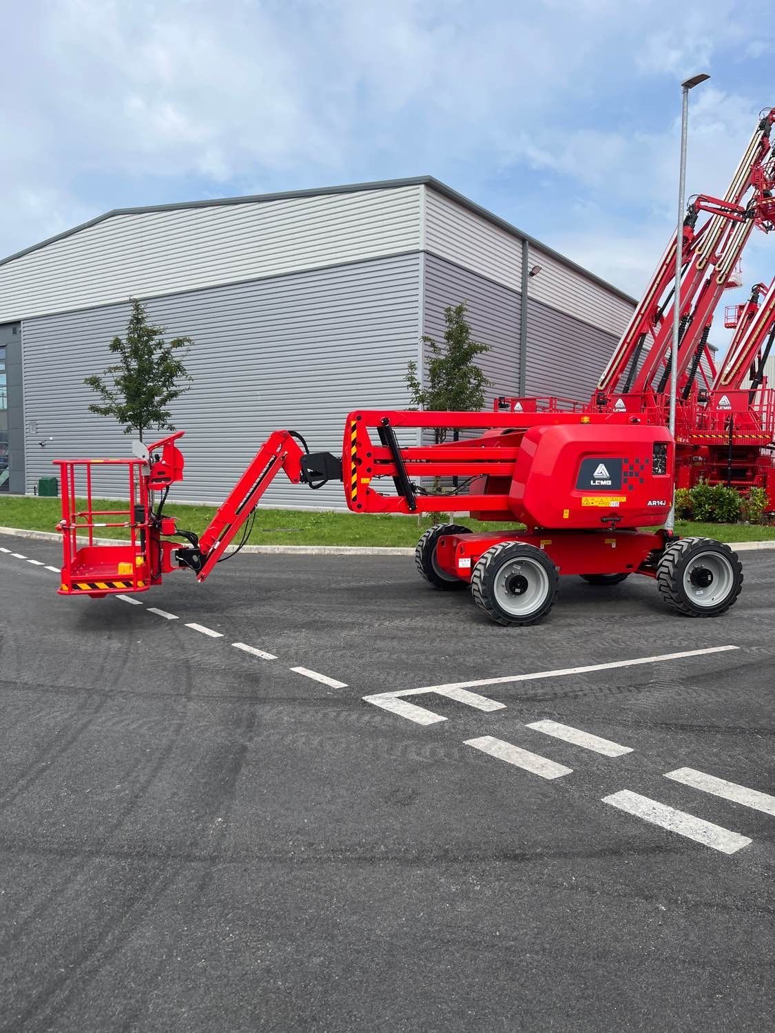 Red aerial work platform on asphalt, boom extended. White building in background.