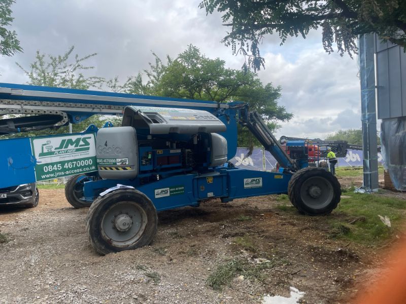 Blue Genie boom lift on construction site, with partially visible building and trees.