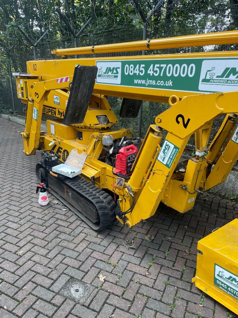 Yellow tracked lift, JMG 25FS, on a brick driveway, next to a wooded area.