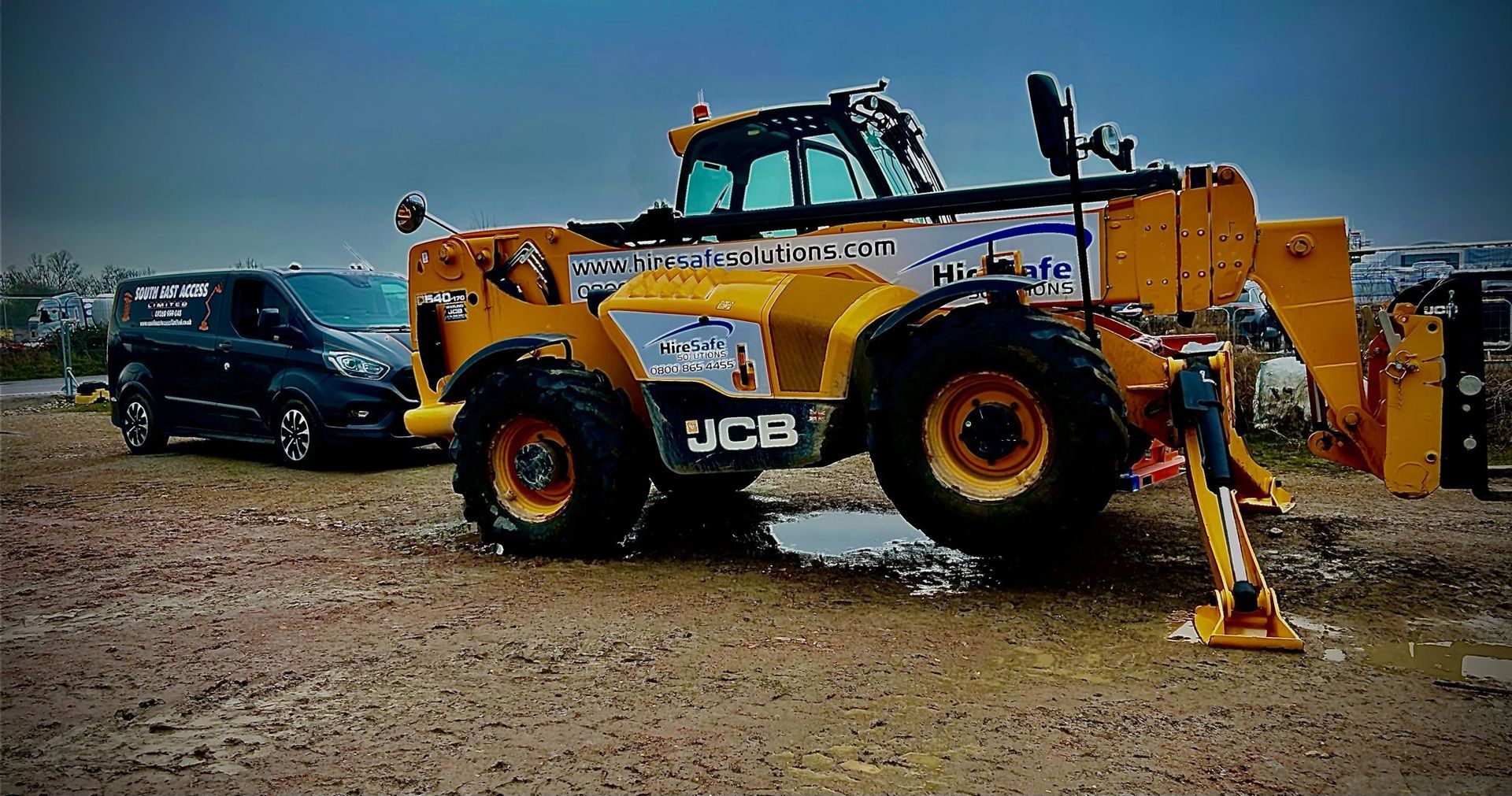 Yellow JCB telehandler on muddy ground next to a dark van. Overcast day.
