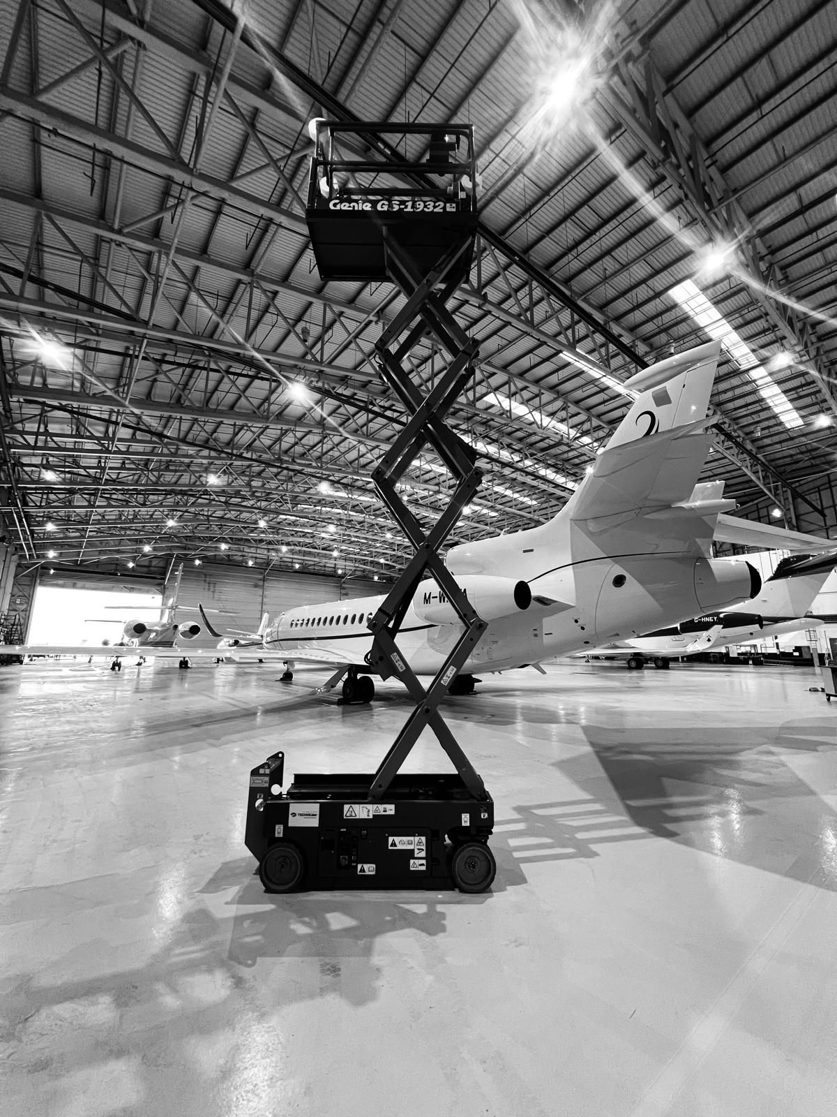 Black and white photo of a scissor lift in an aircraft hangar. A plane is in the background.