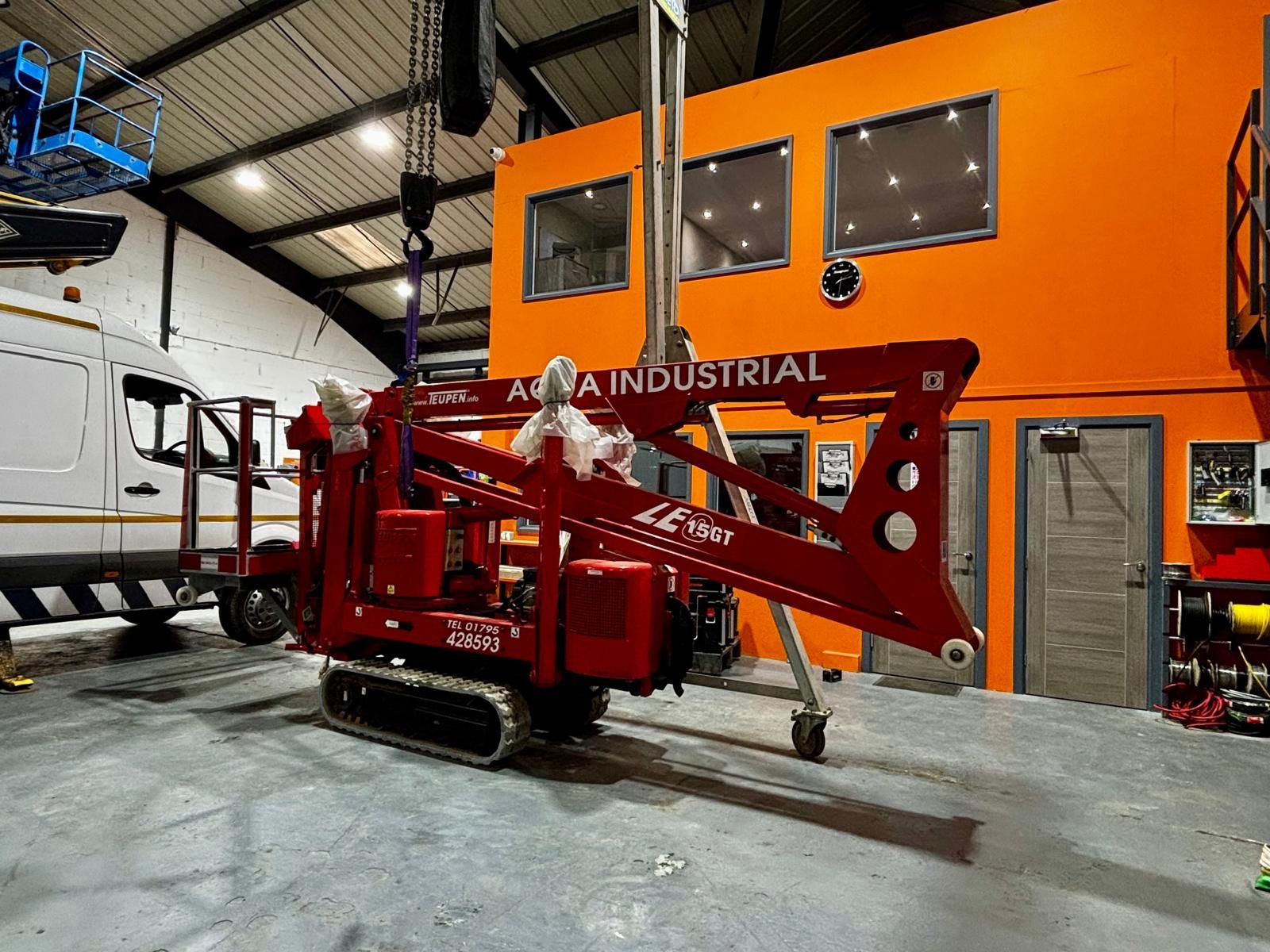 Red lift machine on tracks inside a warehouse. "Aqua Industrial" text is visible on the machine.