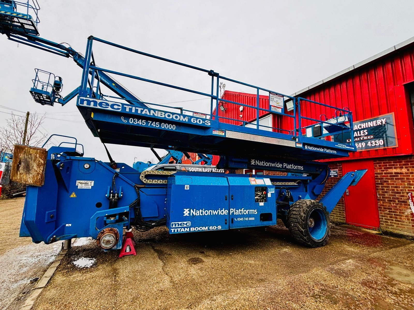 Blue MEC TitanBoom 60-S platform lift on tires, parked next to a red building.