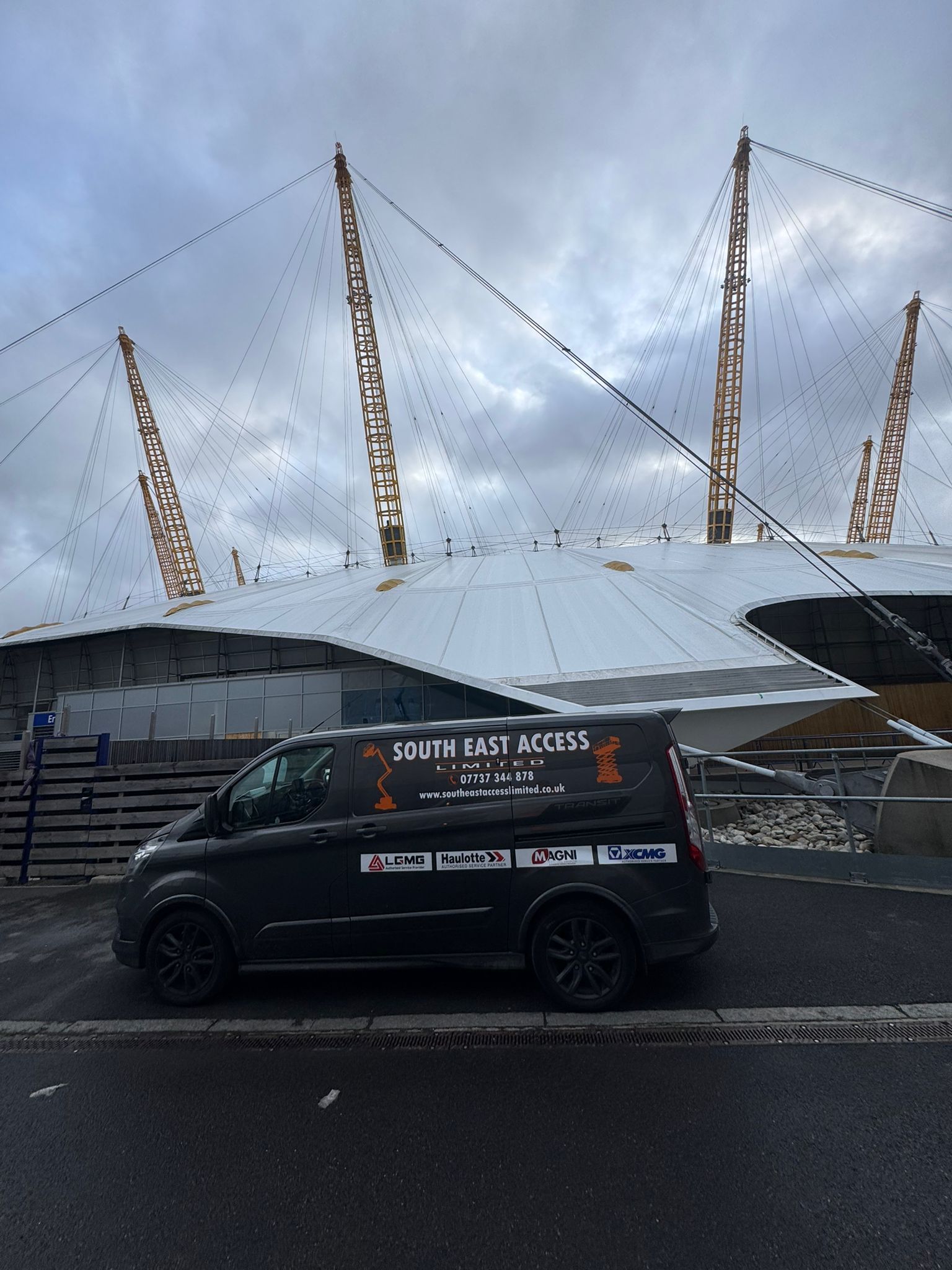 Black van parked in front of The O2 arena, cloudy day. Van has company logo on side.