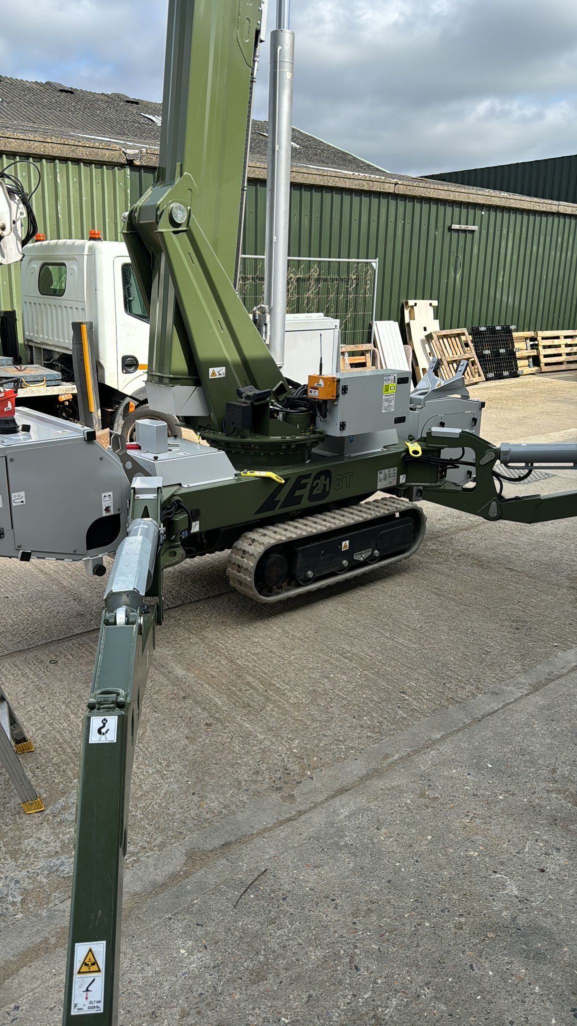 Tracked, olive-green robotic arm in a yard with a truck and pallets visible.