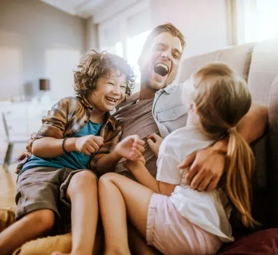 Three people laughing together on a couch in a sunlit living room