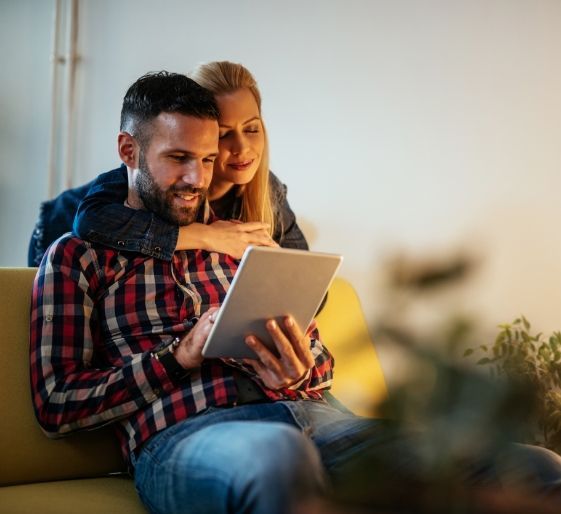 Two people sitting together on a couch, smiling while looking at a tablet.