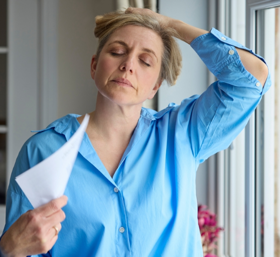 Woman in blue shirt wiping her forehead while holding papers by a window, looking stressed.