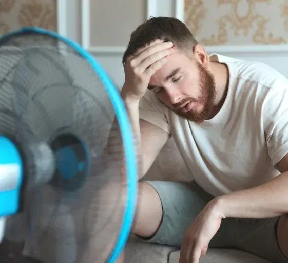 Tired man sitting beside a blue fan, resting his head in his hand indoors