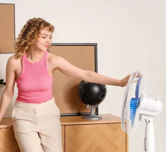 Woman in pink top and beige pants reaching toward a white standing fan in a room