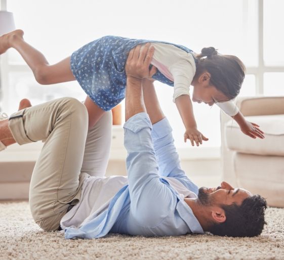 Adult lifting a child indoors on a carpeted floor, both smiling in a bright living room
