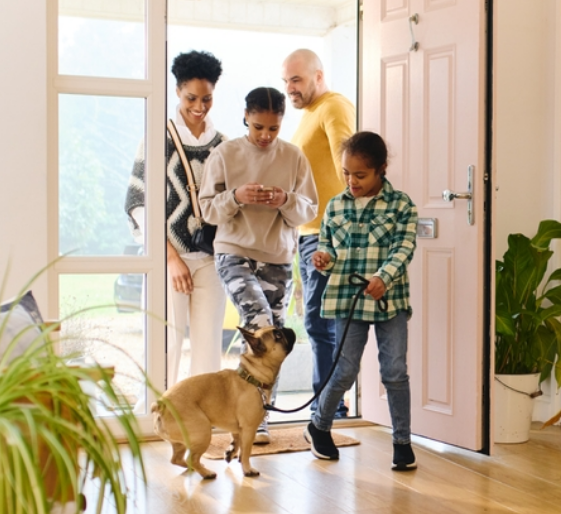 Family standing in a bright doorway, smiling at a small dog on a hardwood floor.