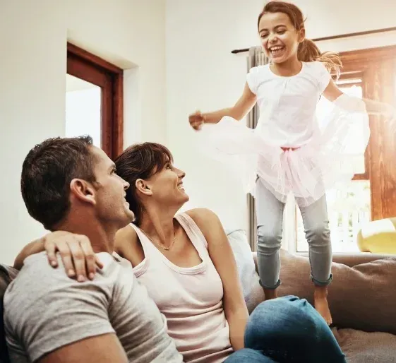 Family smiling on a couch as a child jumps beside them in a sunlit living room