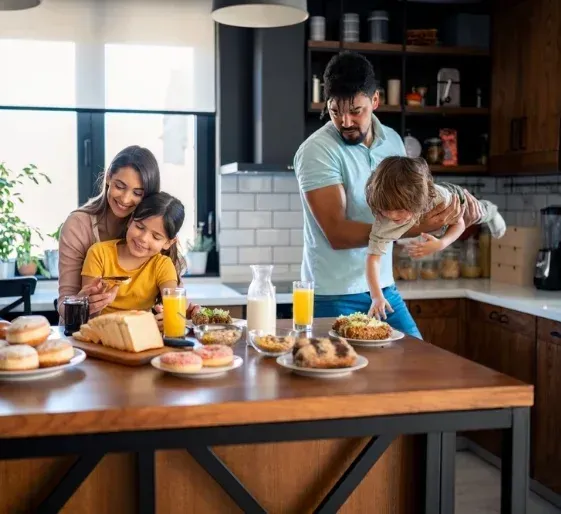 Family preparing breakfast in a modern kitchen, with pancakes, juice, and bread on the table.