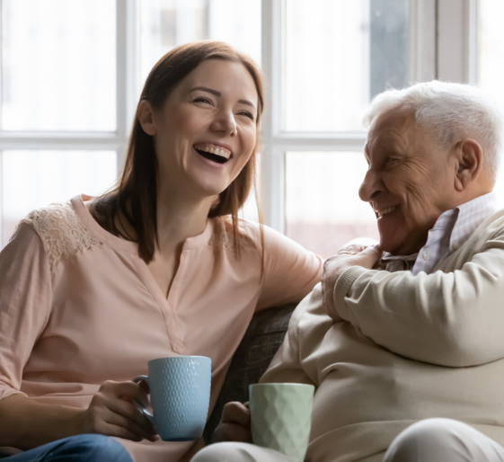 Two people laughing on a couch, holding mugs by a bright window