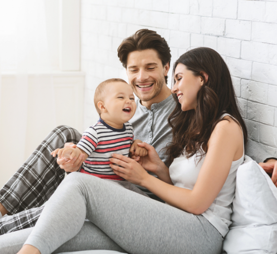 Family sitting on a bed, smiling and playing with a laughing baby in a bright white room