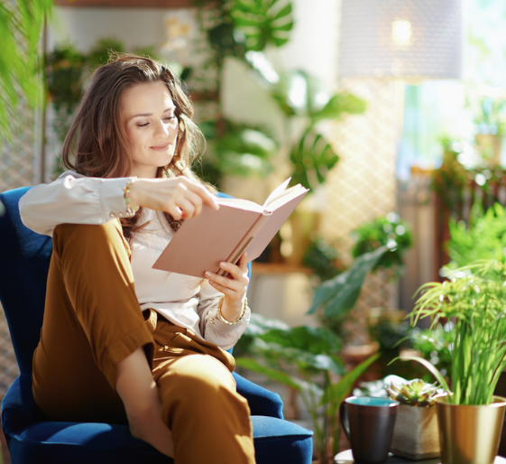 Woman reading a book on a blue chair in a bright room with houseplants and sunlight.
