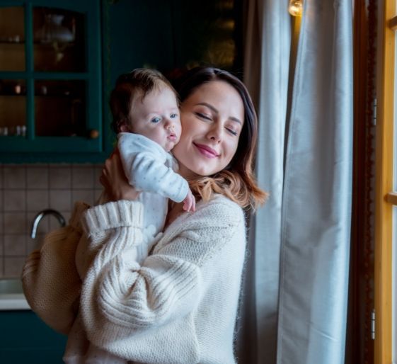 Woman holding a baby by a window, both wearing cozy sweaters, in warm indoor light