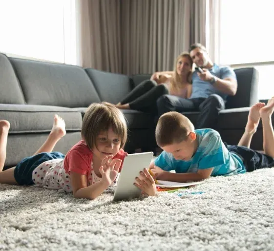 Two children lying on a rug, using a tablet in a living room, with adults sitting on a couch in the background