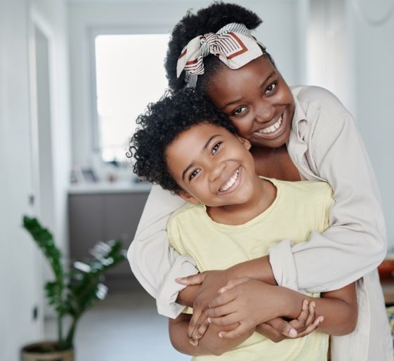 Smiling child hugging an adult indoors, both leaning together in a bright hallway.