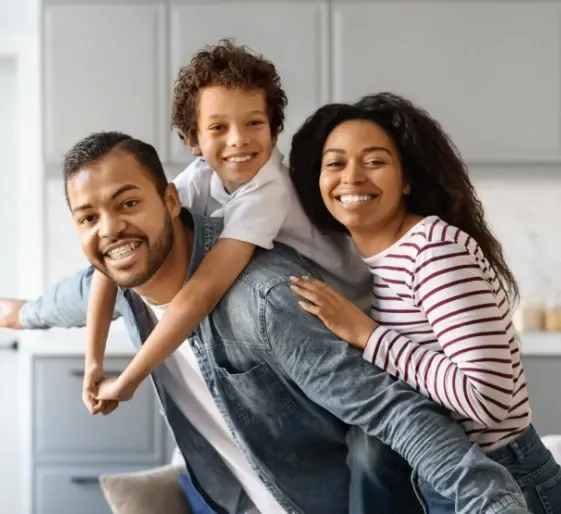 Smiling family posing together indoors, with a child on a parent’s back.