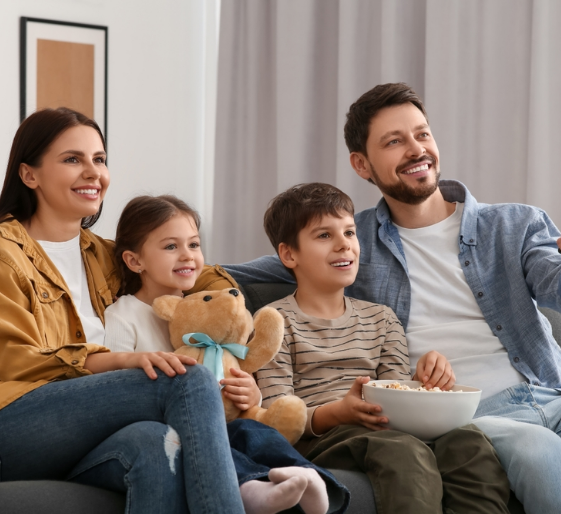 Smiling family on a couch watching TV, with a child holding a teddy bear and another holding a snack bowl