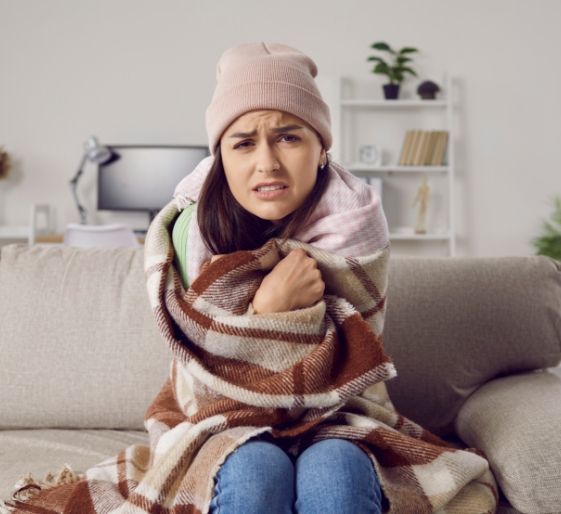 Shivering person on a couch wrapped in a striped blanket, wearing a beanie, in a cozy living room
