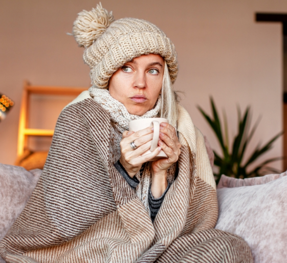 Person wrapped in a blanket and knit hat, holding a mug on a couch in a cozy room