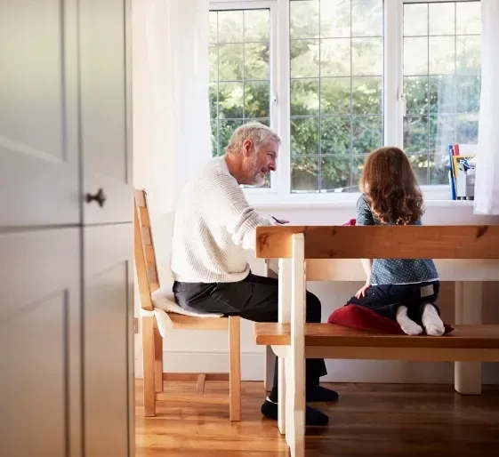 Older man and child seated at a wooden table in a bright kitchen by a window