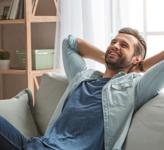 Man relaxing on a couch with hands behind his head, smiling in a bright living room